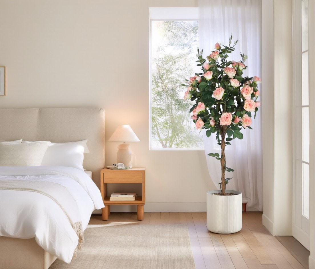 Lichte slaapkamer met wit beddengoed en een linnen hoofdbord. Naast het bed staat een kunstmatige rozenboom van 140 cm met roze bloemen in een witte geribbelde plantenbak, met daglicht door een vitrage erachter.
